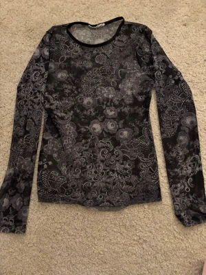 Svart mesh topp med blommigt mönster - Säljer en svart mesh topp med lång ärm och detaljerat blommigt mönster i grått. Toppen är genomskinlig och har en rund halsringning med svart kant. Perfekt att styla lager på lager för en cool look.