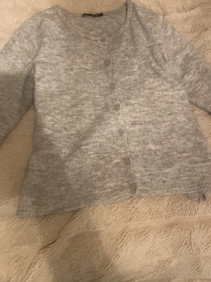 Grå kofta från Lager 157, storlek M - Mysig gråmelerad kofta från 157 i storlek M. Koftan har lång ärm, rund halsringning och knappar hela vägen framtill. Materialet är stickat och känns mjukt mot huden, perfekt för lager-på-lager under kyliga dagar.