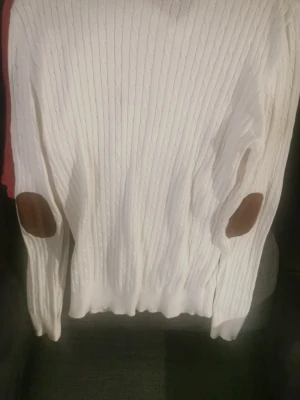 Vit stickad tröja Polo Ralph Lauren - Säljer en vit stickad tröja från Polo Ralph Lauren med v-ringning och brunt broderat logotyp på bröstet. Tröjan har långa ärmar med bruna mockadetaljer på armbågarna och ett klassiskt kabelstickat mönster. Perfekt för höst och vinter!