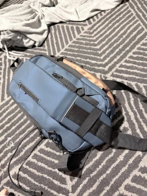 Blå axelväska med USB-port - Säljer en snygg blå axelväska från Fashion med beige detaljer och svarta remmar. Väskan har flera fack med dragkedja, inbyggd USB-port och ett kodlås för extra säkerhet. Perfekt för dig som vill ha med dig tech-prylar och hålla koll på grejerna.