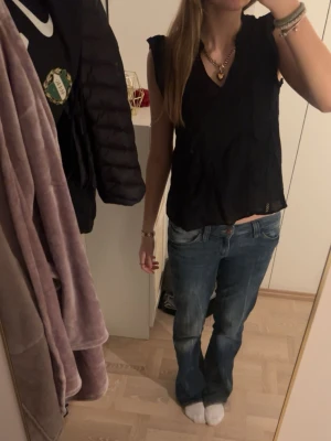 Svart blus från Vero Moda - Svart blus från Vero Moda i storlek XS. Blusen har v-ringning, ärmlös design och fina spetsdetaljer längs framsidan. Tillverkad i ett lätt och luftigt tyg som passar perfekt till jeans eller kjol. Enkel och stilren modell med en feminin touch.