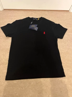 Svart t-shirt från Polo Ralph Lauren - Svart t-shirt från Polo Ralph Lauren med klassisk passform och rund hals. Ikoniska röda loggan broderad på bröstet. Tillverkad i mjuk bomull som känns skön mot huden. Perfekt basic-plagg med stilren vibe.
