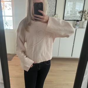 Ljusrosa stickad tröja med rosettdetalj - Superfin ljusrosa stickad tröja med breda ärmar och en söt rosettdetalj framtill. Tröjan har en avslappnad passform och ribbade muddar vid ärmslut och nederkant.