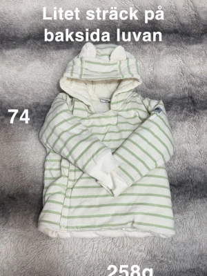 Randig hoodie med öron, stl 74 - Supergullig vit hoodie med ljusgröna ränder och små öron på luvan. Tröjan har knäppning framtill. perfekt för mysiga dagar. Långärmad och med huva för extra komfort.🌸Litet streck på baksidan luvan. Storlek 74