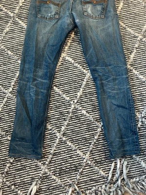 Blå jeans från Nudie Jeans Co W31 L32 - Säljer ett par klassiska blå jeans från Nudie Jeans Co i storlek W31 L32. Jeansen har raka ben, snygga slitningar och kontrastsömmar. De är tillverkade i jeansmaterial och har knäppning med knappar framtill samt bakfickor med dekorativa sömmar.