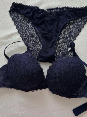 Lindex Mörkblå spets-bh och trosa set S 65B - Snyggt set med mörkblå bh och matchande trosa i spets. Bh:n har vadderade kupor och tunna axelband, medan trosan har fina spetsdetaljer på sidorna och bak. Båda plaggen har ett blommigt spetsmönster som ger en elegant känsla. Bröstvolum 62-65 cm 