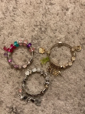 Smyckad silverfärgad charmarmband - Unikt armband i silverfärgad plast med flera utbytbara berlocker som nalle, lås och ankare. Armbandet har coola detaljer som nitar och kubformade pärlor. Perfekt för dig som vill mixa och matcha egna charms och skapa din egen stil.