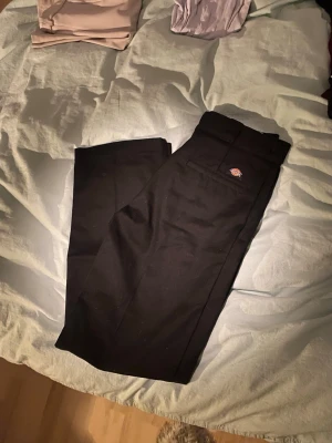 Svarta Dickies 874 byxor W32/L32 - Klassiska svarta Dickies 874 work pants med original fit. Byxorna har raka ben, bälteshällor och Dickies-logga ovanför bakfickan. Tillverkade i slitstarkt material som är perfekt för en clean och tidlös look. Stängs med dragkedja och hake.