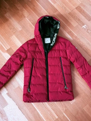 Röd Moncler pufferjacka med huva - Säljer en röd pufferjacka från Moncler med stor huva och svarta detaljer. Jackan har två snedställda dragkedjefickor framtill och tydlig Moncler-logga på huvkanten. Innefickans dragkedja är sönder. Det finns en lite reva på högra axeln. Se bilder. Perfekt för kalla dagar.