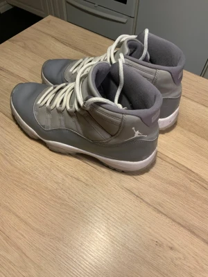 Grå Nike Air Jordan 11 cool grey storlek 45 - Säljer ett par grå Nike Air Jordan 11 sneakers med vita snören och vit sula. Skorna har en hög siluett och den klassiska Jumpman-loggan på hälen. Materialmix av skinn och syntet ger en clean look. Perfekta för dig som gillar streetwear och basketstil.