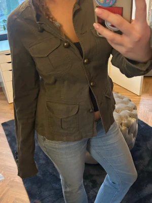Olivgrön utilityjacka med fickor - Säljer en olivgrön utilityjacka med flera stora fickor framtill och snygga knappar. Jackan har en figurnära passform och är tillverkad i ett slitstarkt bomullstyg. Perfekt för dig som gillar en stilren och praktisk look. Insidan syns ej men det är ett rosa camouflage tyg.