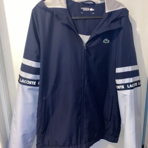 Blå träningsjacka från Lacoste - Säljer en snygg träningsjacka från Lacoste i navy blue och phoenix blue. Jackan har en dragkedja framtill och långa ärmar med vita och blå ränder samt Lacoste-logga på ärmarna. Perfekt för sportiga aktiviteter och våren