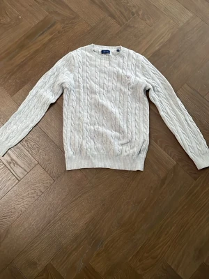 Ljusgrå kabelstickad tröja från Gant - Säljer en ljusgrå kabelstickad tröja från Gant med rund hals och broderad logga på bröstet. Tröjan har långa ärmar och ribbade muddar vid ärmslut och nederkant. Perfekt för kyliga dagar och enkel att matcha med jeans eller kjol.