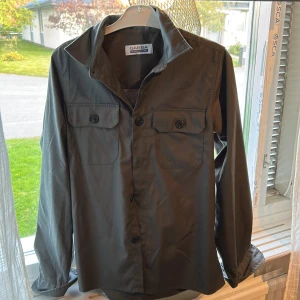 Mörkgrå overshirt från Gabba - Stilren mörkgrå overshirt från Gabba med klassisk krage, två bröstfickor med knappar och långa ärmar. Perfekt lager-på-lager-plagg med snygg passform och diskreta detaljer. Enkel att matcha med det mesta. Storlek s/m 