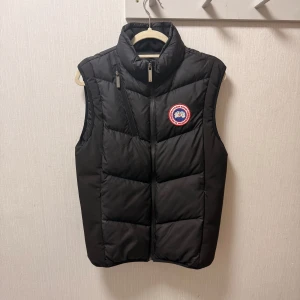 Svart dunväst från Canada Goose - Svart dunväst från Canada Goose med hög krage, dragkedja och klassisk logotyp på bröstet. Västen har quiltad design och är perfekt för lager-på-lager. Snygg och stilren look med diskret Canada Goose-märke på ryggen. Strlk S men passar M