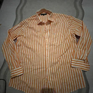 Randig orange skjorta från Dahlin XXL -                                 Snabb leverans.                                                              Snygg skjorta från Dahlin i storlek XXL med vertikala orange och vita ränder. Klassisk krage, lång ärm och bröstficka med liten logotyp. Skjortan har knäppning framtill och är tillverkad i mjuk bomull, perfekt för dig som gillar färg och mönster.