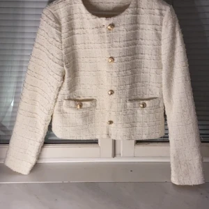Vit kavajjacka med guldknappar - ‼️KAN SÄNKAS‼️Säljer en stilren vit kavajjacka i strukturerat tyg med snygga guldknappar framtill och vid fickorna. Jackan har rund halsringning, raka axlar och är midjekort. Har använts 2 gånger tidigare.