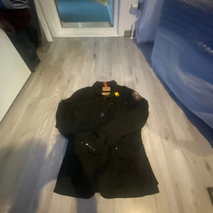 Svart Parajumpers jacka slim fit S - Säljer en svart jacka från Parajumpers i slim fit-modell, storlek S. Jackan har klassisk krage, flera fickor framtill, snygga knappar och en gul detalj vid halsen. Märkespatch på ärmen och orange hängare i nacken. Perfekt för dig som gillar stilren och funktionell design.