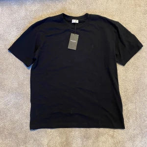 Saint Laurent Relaxed Fit Tshirt  - Svart t-shirt från Saint Laurent med diskret YSL-broderi på bröstet. Klassisk rund halsringning och korta ärmar. Tillverkad i mjuk bomull för en clean och stilren look. Perfekt för dig som gillar minimalistisk design och premiumkänsla.