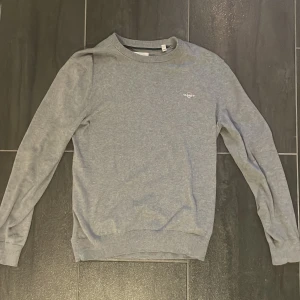 Grå sweatshirt från GANT - En stilren grå sweatshirt från GANT med broderad logga på bröstet. Tröjan har rund halsringning, långa ärmar och ribbade muddar vid ärmslut och nederkant. Perfekt för dig som gillar en clean och klassisk look.