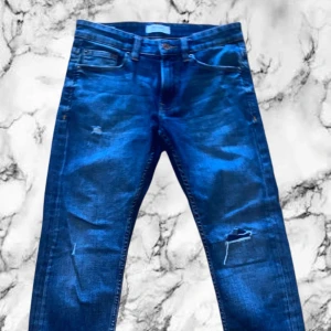 Blå skinny jeans med slitningar - Säljer ett par blå skinny jeans med coola slitningar på benen. Jeansen har klassisk femficksdesign, smal passform och är tillverkade i stretchigt denim för extra komfort. Perfekta för dig som gillar en trendig och avslappnad look.