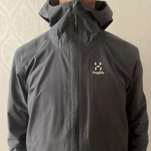 Grå vindjacka från Haglöfs med huva - Säljer en grå vindjacka från Haglöfs med justerbar huva och hel dragkedja framtill. Jackan är tillverkad i slitstark polyester och har meshfoder på insidan för extra komfort. Perfekt för blåsiga dagar och outdoor-aktiviteter. Snygg och funktionell design.