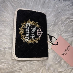 Juicy Couture svart och beige plånbok - Plånbok från Juicy Couture i svart sammet med quiltad framsida, broderad logga, gulddetaljer och beige kanter. Insidan har flera kortfack och tryckknapp. Baksidan är beige med dragkedjefack och kedjedetalj. Perfekt för dig som gillar lyxig och trendig stil.