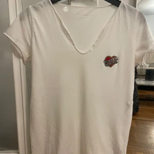 Vit t-shirt Zadig & Voltaire hjärtdetalj - Vit t-shirt från Zadig & Voltaire med v-ringning och små knappar vid halsen. På bröstet finns en broderad hjärtpatch med märkesnamnet. T-shirten är kortärmad och i mjuk bomull, perfekt för en avslappnad och cool look.