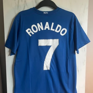 Blå Manchester United Ronaldo tröja - Säljer en blå Manchester United fotbollströja från Nike med Ronaldo och nummer 7 på ryggen. Tröjan har korta ärmar, AIG som sponsor på bröstet och klubbmärket broderat. Materialet är lätt och andas, perfekt för match eller träning.