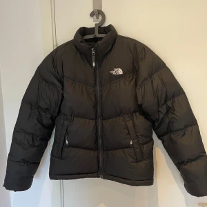 Svart pufferjacka från The North Face - Säljer en svart pufferjacka från The North Face i nyskick. Köpt på Stadium förra året och endast använd några ggr. Jackan har hög krage och dragkedja framtill, och  två fickor med dragkedja och klassisk logga broderad på bröstet och ryggen. Perfekt för kalla dagar och riktigt snygg till streetwear.