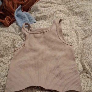 Ljusrosa ribbad linnetopp Iconic - Supersöt ljusrosa ribbad linnetopp från Iconic. Toppen är ärmlös med klassisk rak halsringning och har en liten broderad detalj nertill. Perfekt för varma dagar och enkel att matcha med jeans eller shorts.