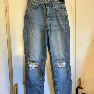 Blå ripped jeans från Perfect Jeans/Gina Tricot - Säljer ett par ljusblå jeans från Perfect Jeans med raka ben och slitna hål på båda knäna. Klassisk femficksmodell med dragkedja och knapp i midjan. Jeansen har en avslappnad passform och är tillverkade i bomull.