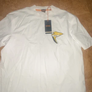 Vit BOSS t-shirt med gul detalj - Vit t-shirt från BOSS i relaxed fit med coolt gult och svart märke på bröstet där det står 'BRT 3055'. Klassisk rund hals och korta ärmar. Materialet är mjuk bomull och passar perfekt för dig som gillar stilrena plagg med en twist.