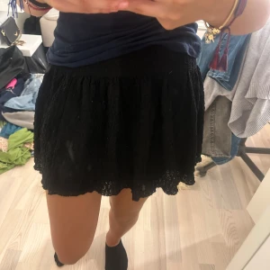 Svart spetsig kortkjol - Supersöt svart kortkjol med volangig och spetsig struktur. Kjolen har en lätt utställd form och resår i midjan för skön passform. Perfekt att styla med en enkel topp eller stickad tröja.