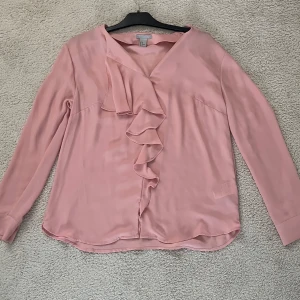 Ljusrosa blus med volang  - Säljer en ljusrosa blus med långa ärmar och v-ringning. Framsidan har en snygg volangdetalj som ger en extra touch. Blusen är tillverkad i ett mjukt, lätt material och har en feminin, flowy passform. Perfekt för dig som gillar stilrena och romantiska plagg.