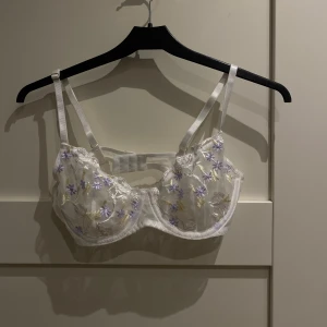 Vit blommig bh - Superfin vit korsett-bh med broderade blommor i lila och gult. Bh:n har justerbara axelband och hakknäppning bak. 