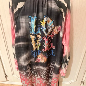 Färgstark tie-dye klänning med tryck - Unik långärmad klänning i tie-dye med rosa, svart och vita toner. Framsidan har knappar vid halsen och baksidan är dekorerad med färgglatt 'LOVE FOR ALL'-tryck och fjärilar. Lätt och flowy modell, perfekt för dig som gillar statement-plagg.  Love forever