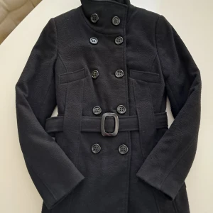 Svart kappa från Staroma i kashmir/ull - Stilren svart kappa från Staroma i kashmir- och ullmix. Dubbelknäppt design med hög krage, bälte i midjan och snygga knappar framtill. Perfekt för dig som vill ha en klassisk och tidlös look under kyliga dagar. (Tjock foder) 