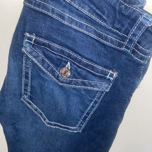 Blå bootcut jeans från Gina Tricot - Snygga blå jeans från Gina Tricot i storlek 164. Modellen har bootcut-ben, markerade vita sömmar och fickor med knappdetaljer bak. Klassisk femficksdesign och gylf med knapp. Perfekta för dig som gillar en retroinspirerad look.