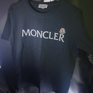 Svart Moncler t-shirt med logga - Svart t-shirt från Moncler med stor vit logotyp framtill och klassisk rund hals. T-shirten har korta ärmar och en broderad Moncler-patch på bröstet. Tillverkad i mjuk bomull för en skön känsla. Perfekt för dig som gillar stilrena och exklusiva plagg.