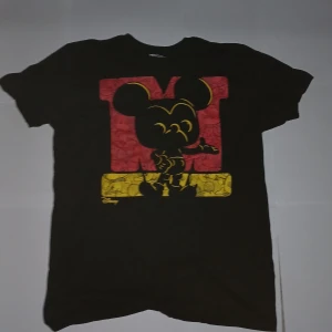 Svart Disney t-shirt med Mickey Mouse - Svart t-shirt från Funko x Disney med stor färgglad Mickey Mouse-tryck i rött och gult på bröstet. Klassisk passform och rund halsringning. Tillverkad i 100% bomull, perfekt för dig som älskar Disney och vill sticka ut med en ikonisk look.