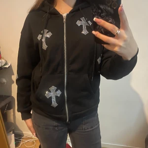 Svart hoodie med strasskors och dragkedja - Svart hoodie med huva och dragkedja framtill, dekorerad med flera glittriga strasskors på bröstet, ärmen och fickan. Mjuk bomullskänsla och snörning i huvan.