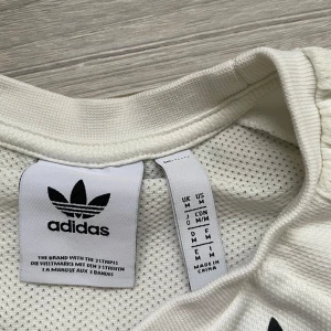 Vit Adidas t-shirt med meshdetaljer - Vit t-shirt från Adidas med klassiska tre ränder på axlarna och svart logga på bröstet. Tillverkad i luftig polyester med meshpaneler för extra ventilation. Perfekt för dig som gillar sportig och clean stil.