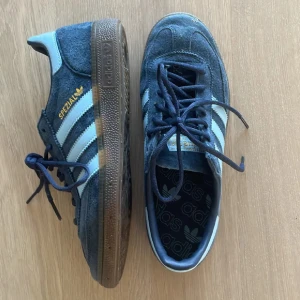Adidas Spezial blå sneakers - Säljer ett par Adidas Spezial sneakers i mörkblå mocka med ljusblå detaljer och klassiska vita ränder på sidorna. Skorna har snörning, gummisula och guldfärgad Spezial-logga. Perfekta för dig som gillar retrostil och streetwear.