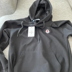 Svart hoodie från Moncler med huva - Svart hoodie från Moncler med klassisk huva och dragsko, samt broderad logga på bröstet. Tröjan har en stor magficka och är tillverkad i mjuk bomull. Perfekt för dig som gillar stilrena och exklusiva plagg.