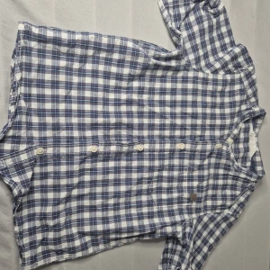 Blårutig henley-skjorta från H&M - Snygg blårutig henley-skjorta från H&M i bomull. Skjortan har korta ärmar, knappar framtill och en liten bröstficka. Perfekt för dig som gillar en avslappnad men ändå stilren look.