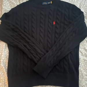 Svart kabelstickad tröja Polo Ralph Lauren - Svart kabelstickad tröja från Polo Ralph Lauren med klassisk röd broderad logga på bröstet. Tröjan har rund hals, ribbade muddar och lång ärm. Perfekt för dig som gillar stilrena och tidlösa plagg med premiumkänsla.