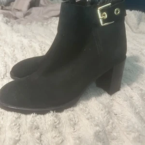 Svarta mockaboots Tommy Hilfiger - Snygga svarta boots från Tommy Hilfiger i mocka med chunky klack och guldfärgade spänndetaljer upptill. Dragkedja på sidan och rund tå. Insidan har ett mönstrat foder med stjärnor i rött, vitt och blått. Perfekta för dig som vill ha en stilren men cool look.