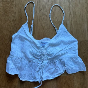 Vit linnetopp med volang från Bershka - Supersöt vit linnetopp från Bershka med smala axelband, volangkant nertill och snygg snörning framtill. Toppen är croppad och har en lätt rynkad effekt över bysten. 