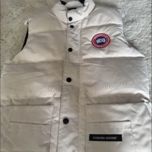 Vit dunväst från Canada Goose XS - Snygg vit dunväst från Canada Goose med klassisk logga på bröstet och svarta knappar. Västen har två stora fickor framtill och dragkedja under knappslån. Perfekt för lager-på-lager och riktigt varm med dunfyllning.